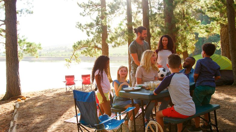 Two Familes Having Picnic Camping By Lake On Hiking Adventure In Forest