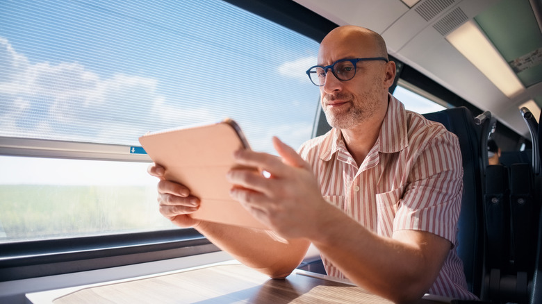 Bald man wearing glasses reading on a digital tablet seated by a train window