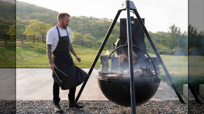 A chef holding tongs and watching an outdoor smoker outside the Switchback Restaurant at Cataloochee Ranch in Maggie Valley, North Carolina.