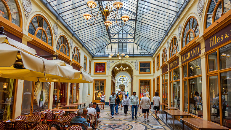 Pedestrians strolling beneath Galerie Vivienne's glass ceiling, past glass storefronts and café tables