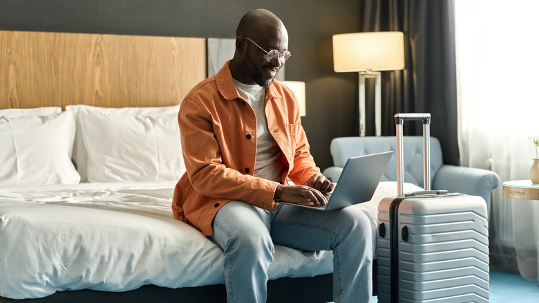 A man sitting on a hotel room bed and looking at a laptop while a suitcase sits nearby